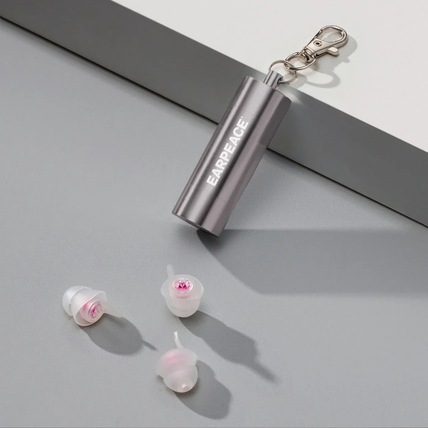 Three clear EARPEACE MUSIC PRO earplugs with pink filters beside the metal carry case.